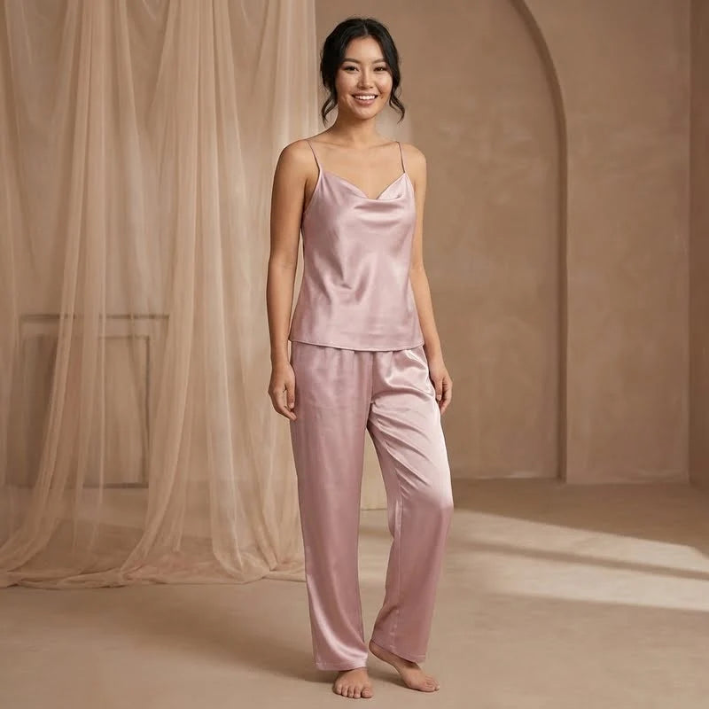 Femme souriante portant un pyjama en satin rose à fines bretelles avec pantalon fluide, debout dans un décor beige élégant avec voilage léger, style photo e-commerce douce et raffinée.