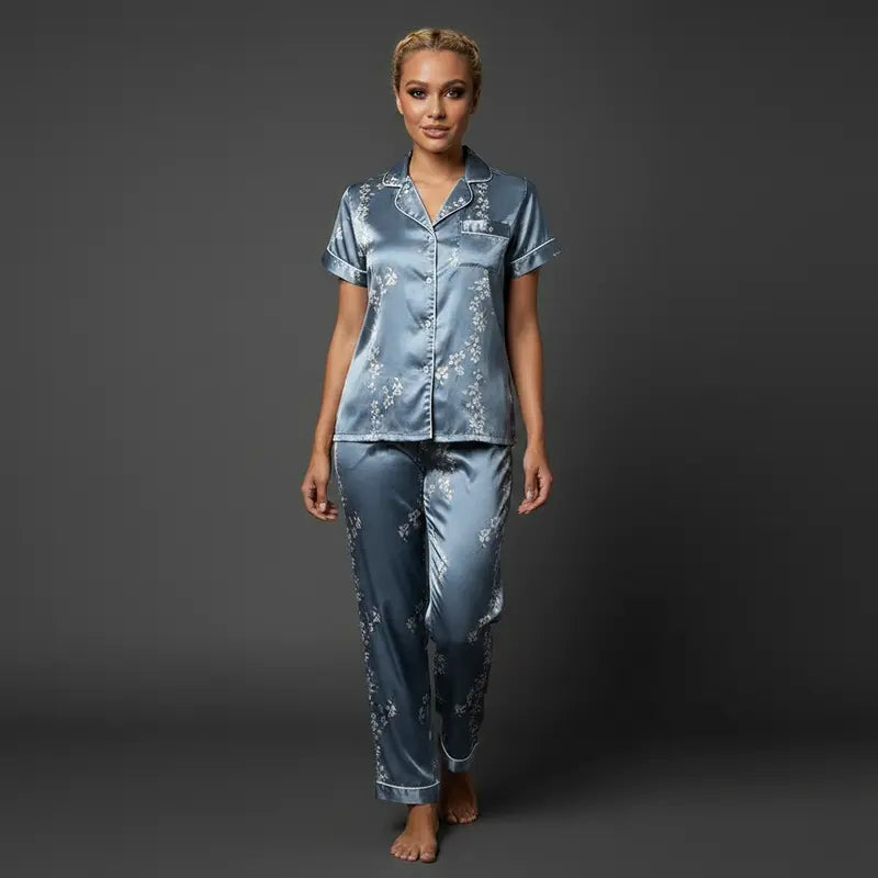 Femme portant un pyjama en satin bleu clair avec imprimé floral, composé d’une chemise boutonnée à manches courtes et d’un pantalon long, debout face à la caméra sur fond gris foncé.