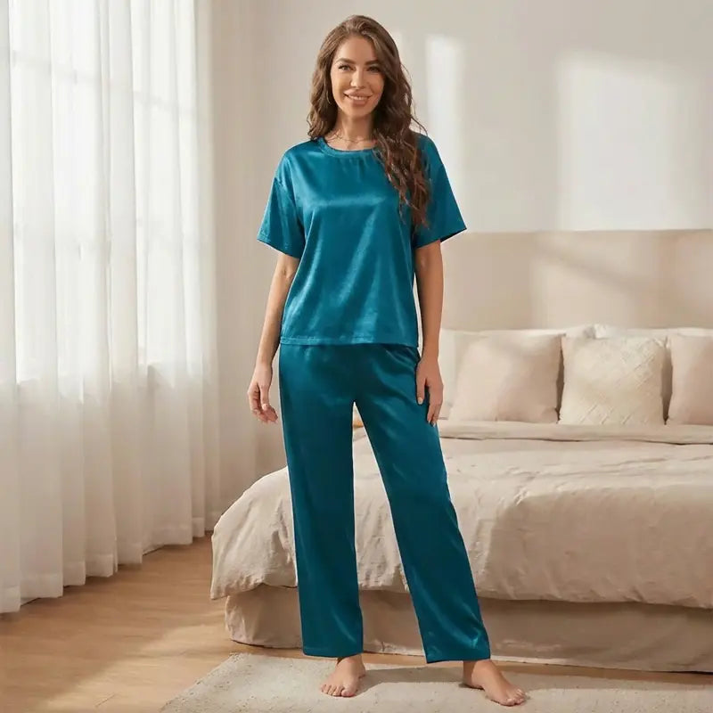 Femme souriante portant un pyjama en satin vert à manches courtes avec pantalon assorti, debout dans une chambre lumineuse avec lit en arrière-plan, style photo e-commerce naturel et confortable.