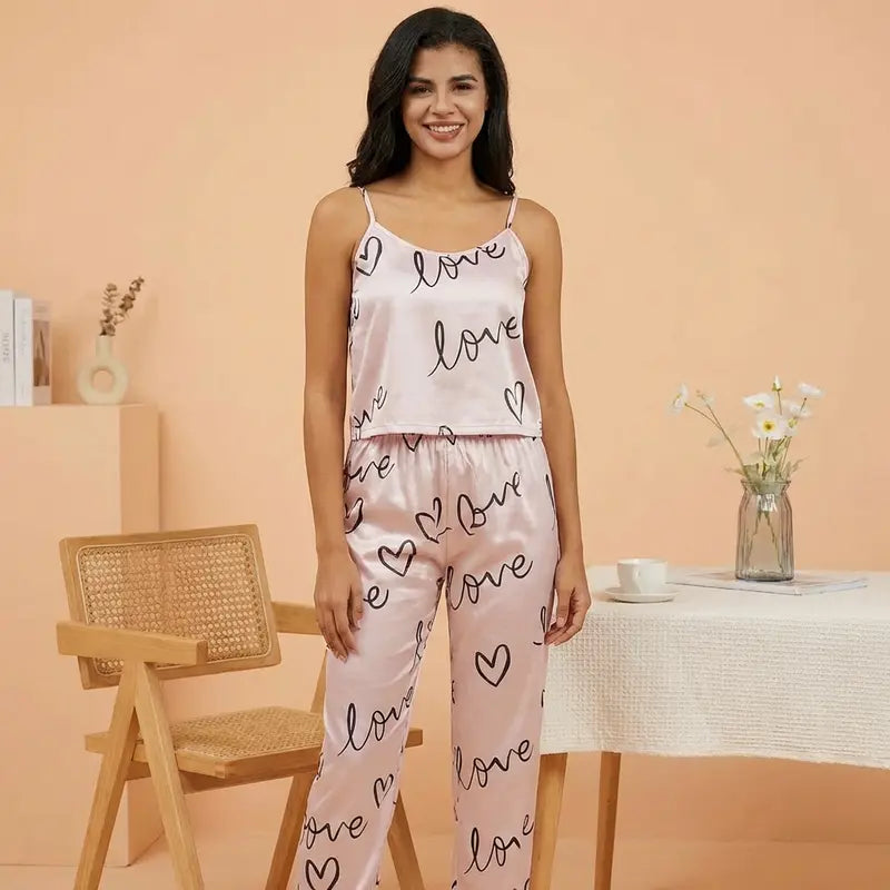 Femme souriante portant un pyjama en satin rose clair à fines bretelles avec motifs cœurs et inscription « love », debout dans un intérieur lumineux au style doux et minimaliste.