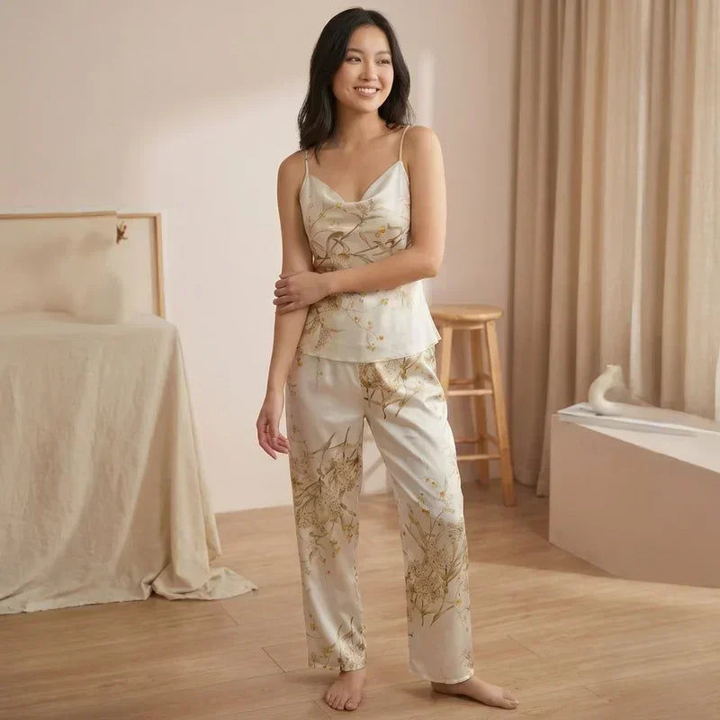 Femme souriante portant un pyjama en satin beige clair à fines bretelles avec motifs floraux délicats et pantalon assorti, posant dans une chambre lumineuse au style naturel et épuré.