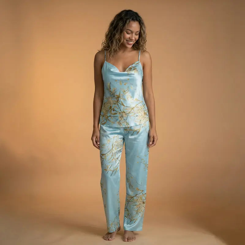 Femme souriante portant un pyjama en satin bleu clair à fines bretelles avec motifs floraux dorés et pantalon assorti, posant sur fond beige, style e-commerce élégant et raffiné.