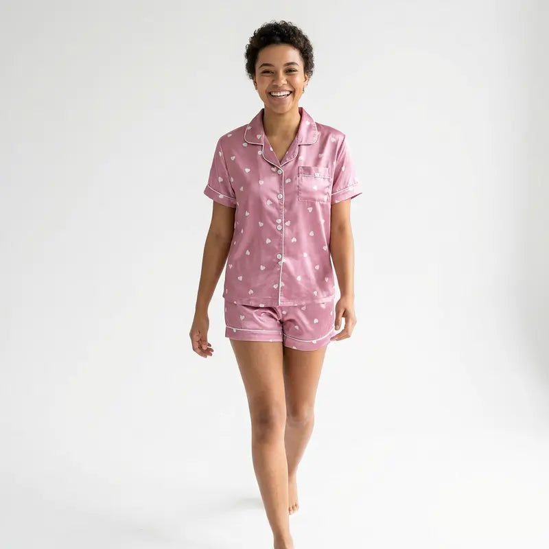 Femme souriante portant un pyjama satin rose à manches courtes avec short assorti et motifs cœurs, debout sur fond neutre, style e-commerce épuré.