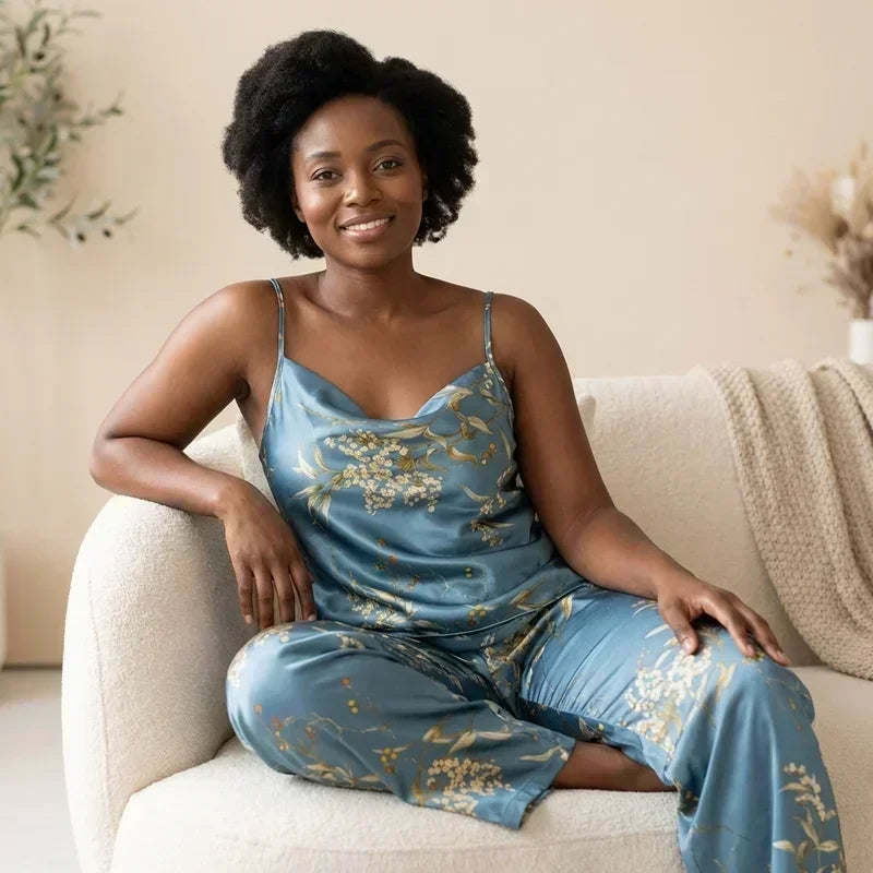 Femme souriante assise sur un fauteuil portant un pyjama en satin bleu à fines bretelles avec motifs floraux dorés et pantalon assorti, dans un intérieur chaleureux et lumineux, style e-commerce élégant et confortable.